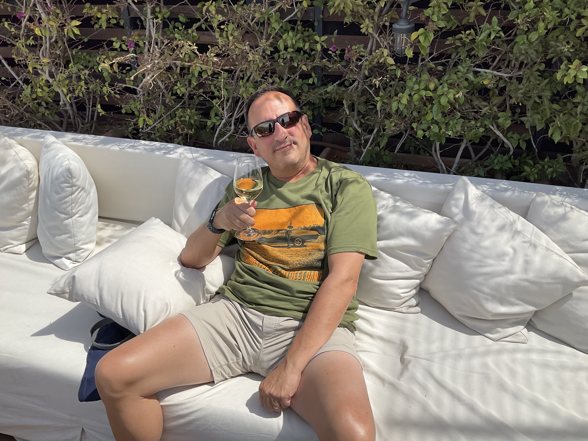 He-Stache in sunglasses lounging on a white outdoor sofa, holding a drink