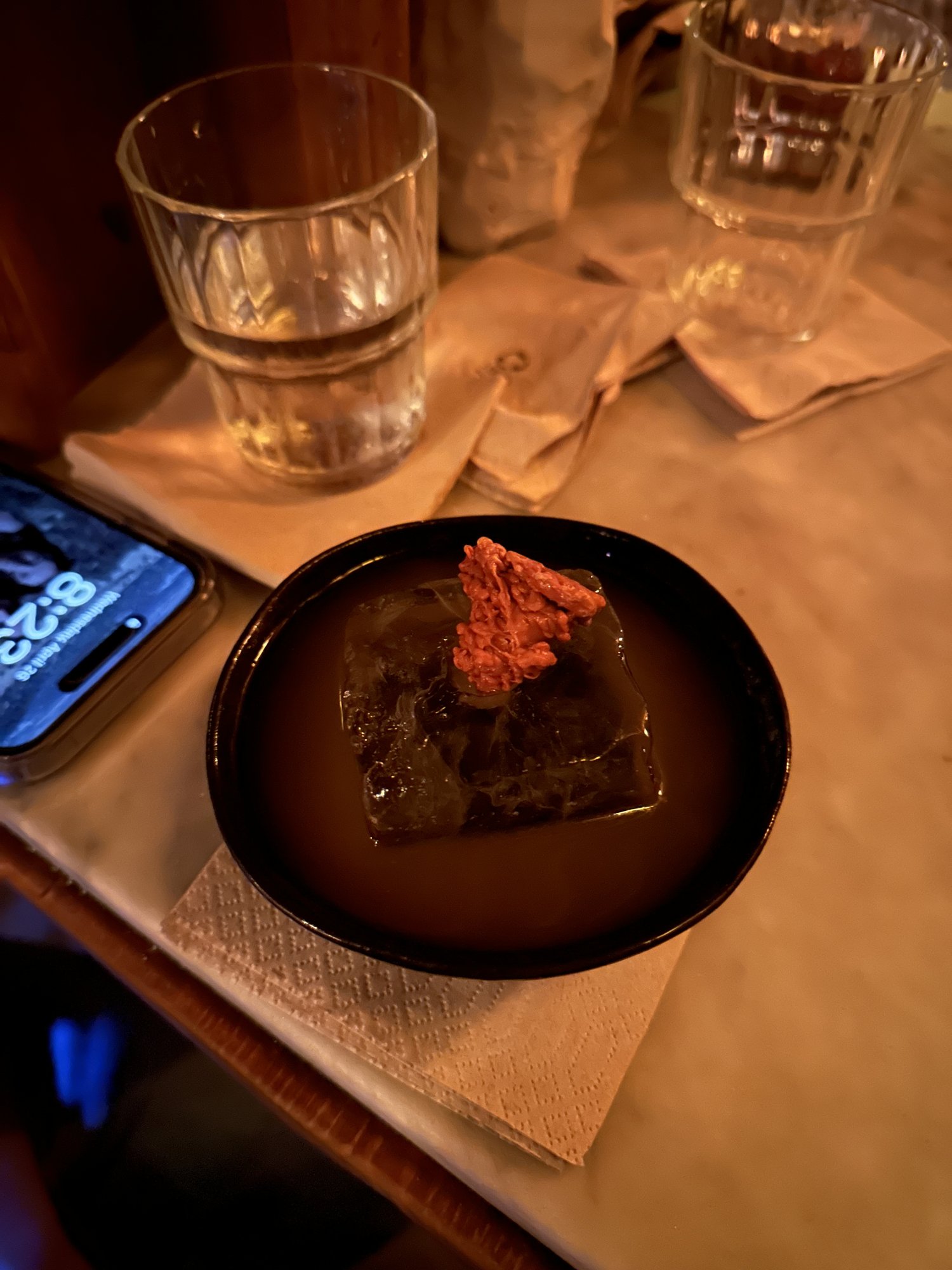 A small dark bowl holding one bite with a red garnish, beside empty cocktail glasses