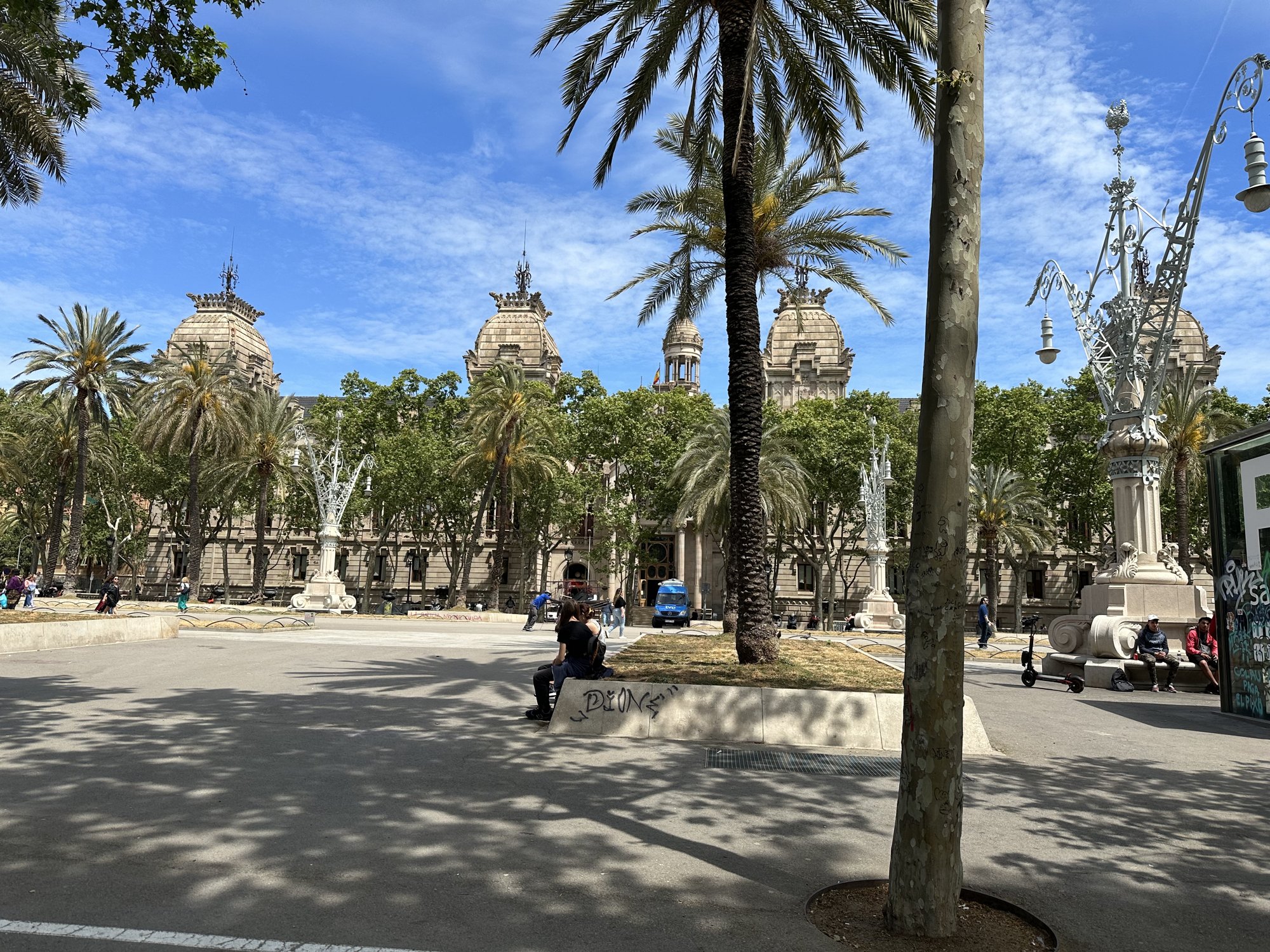 A wide palm-lined promenade with benches under a blue sky, with a large ornate building with towers behind