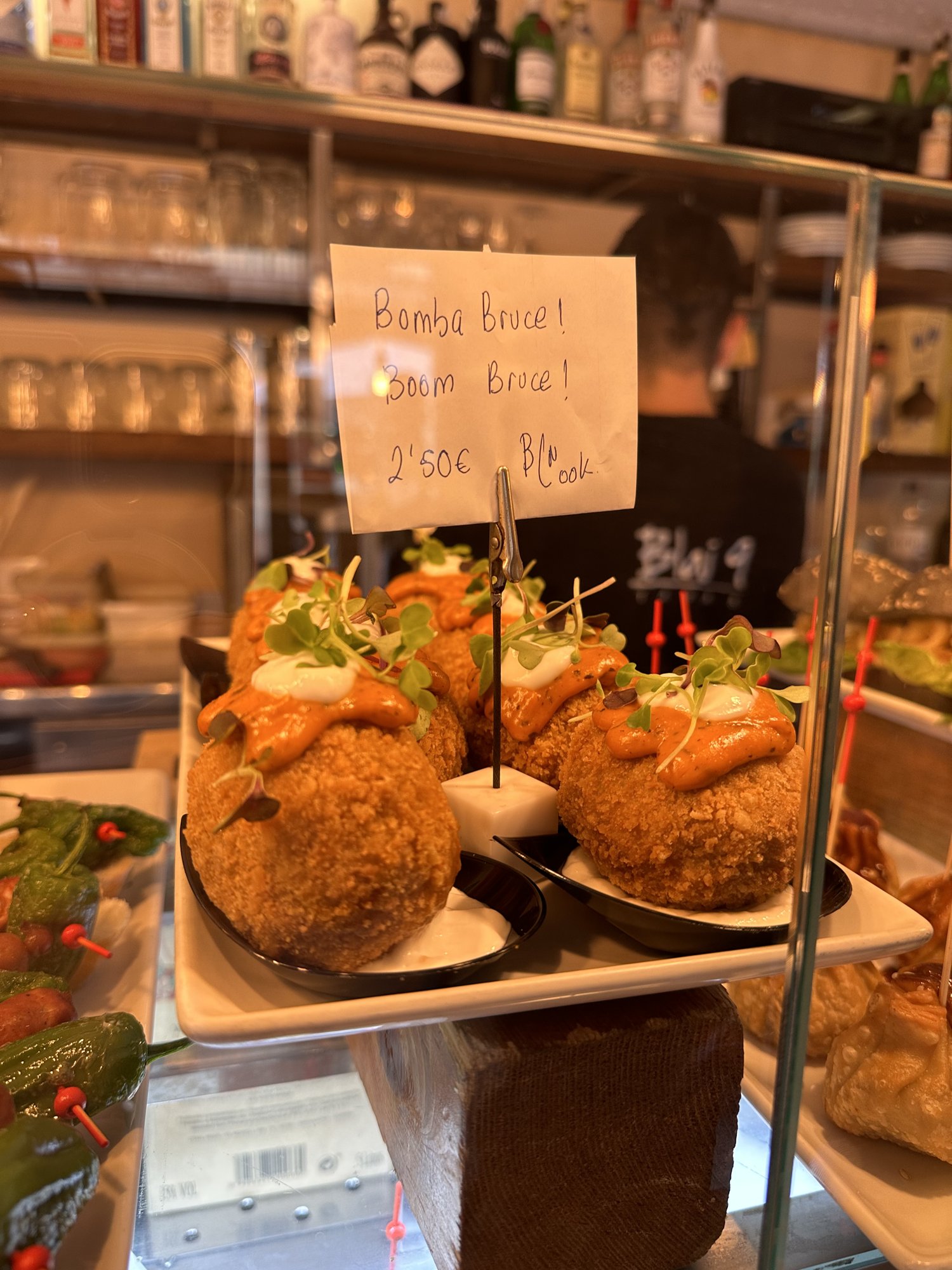 A counter display of fried bombas topped with sauce, with a handwritten sign behind