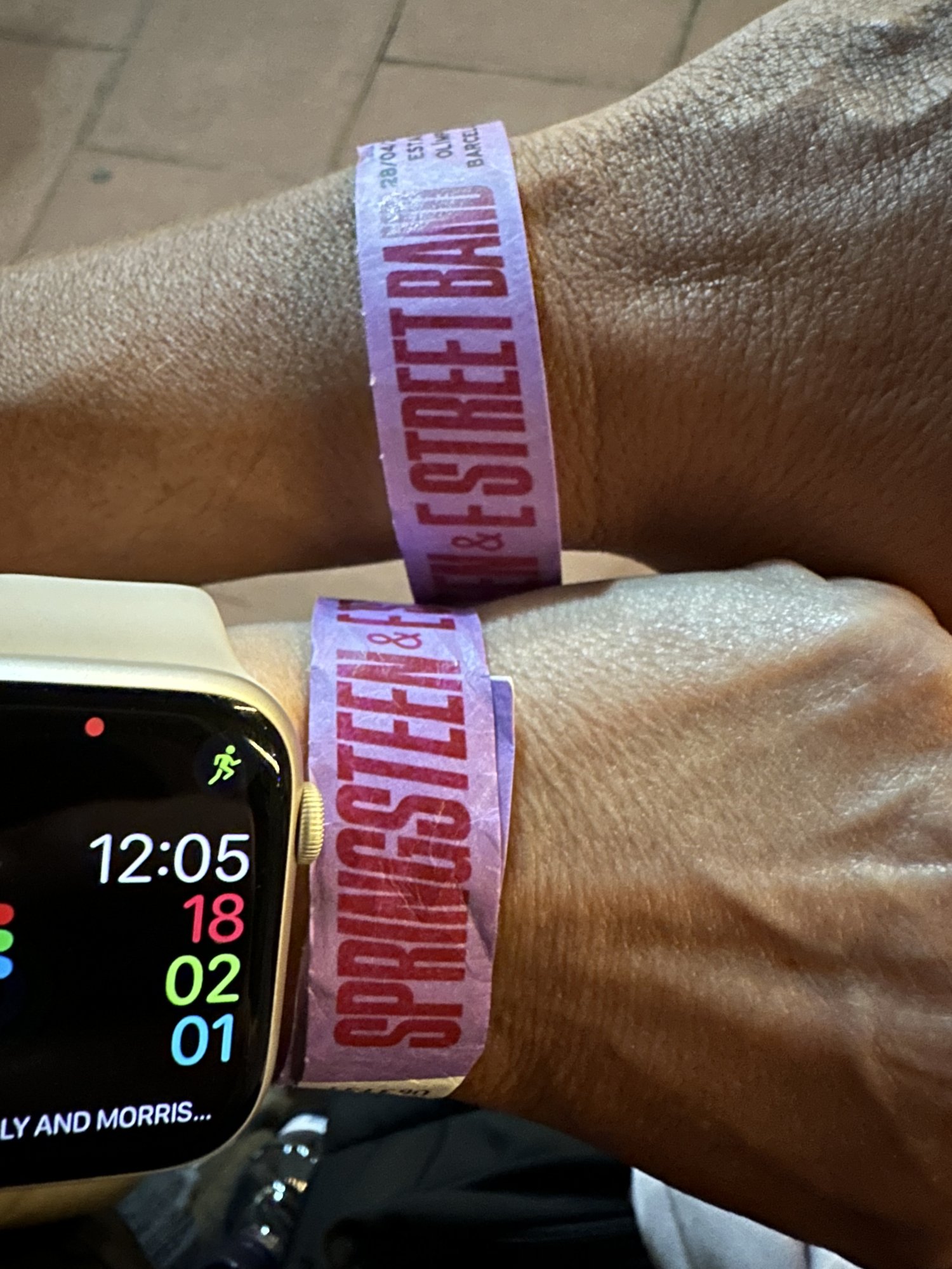 A wrist wearing a Springsteen and E Street Band wristband next to a smartwatch showing 12:05