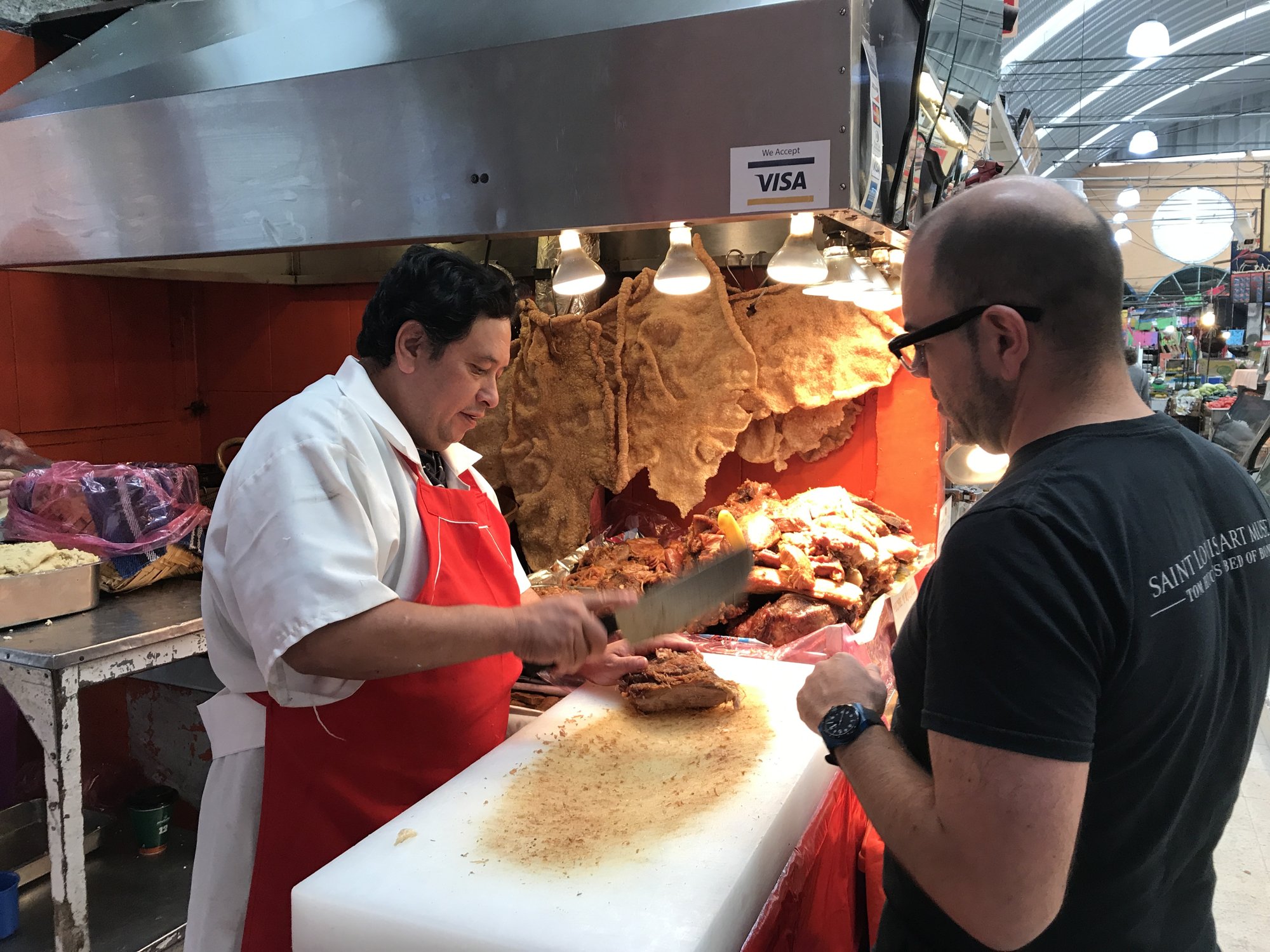 A vendor in a red apron carving meat at a market counter