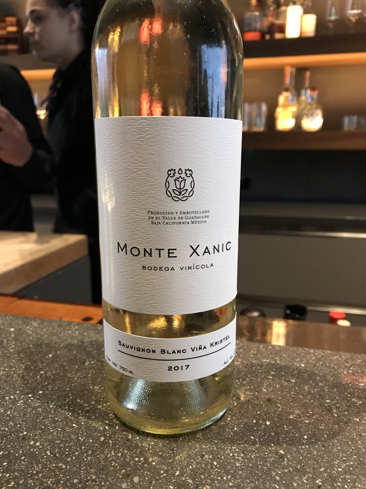 A bottle of Monte Xanic white wine on a dark bar beside wine glasses