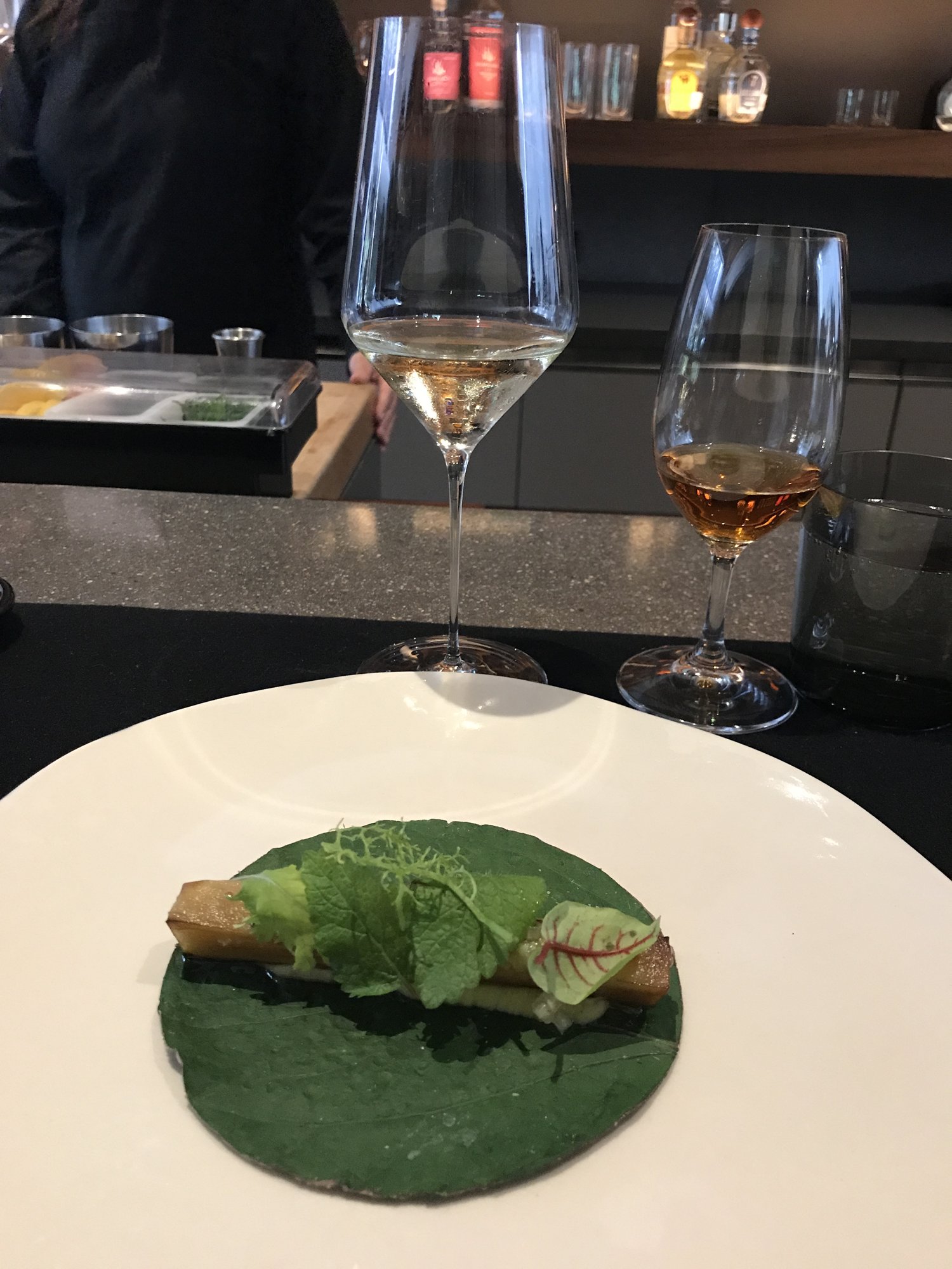 A taco wrapped in a green leaf on a white plate at a dark bar, wine glasses behind