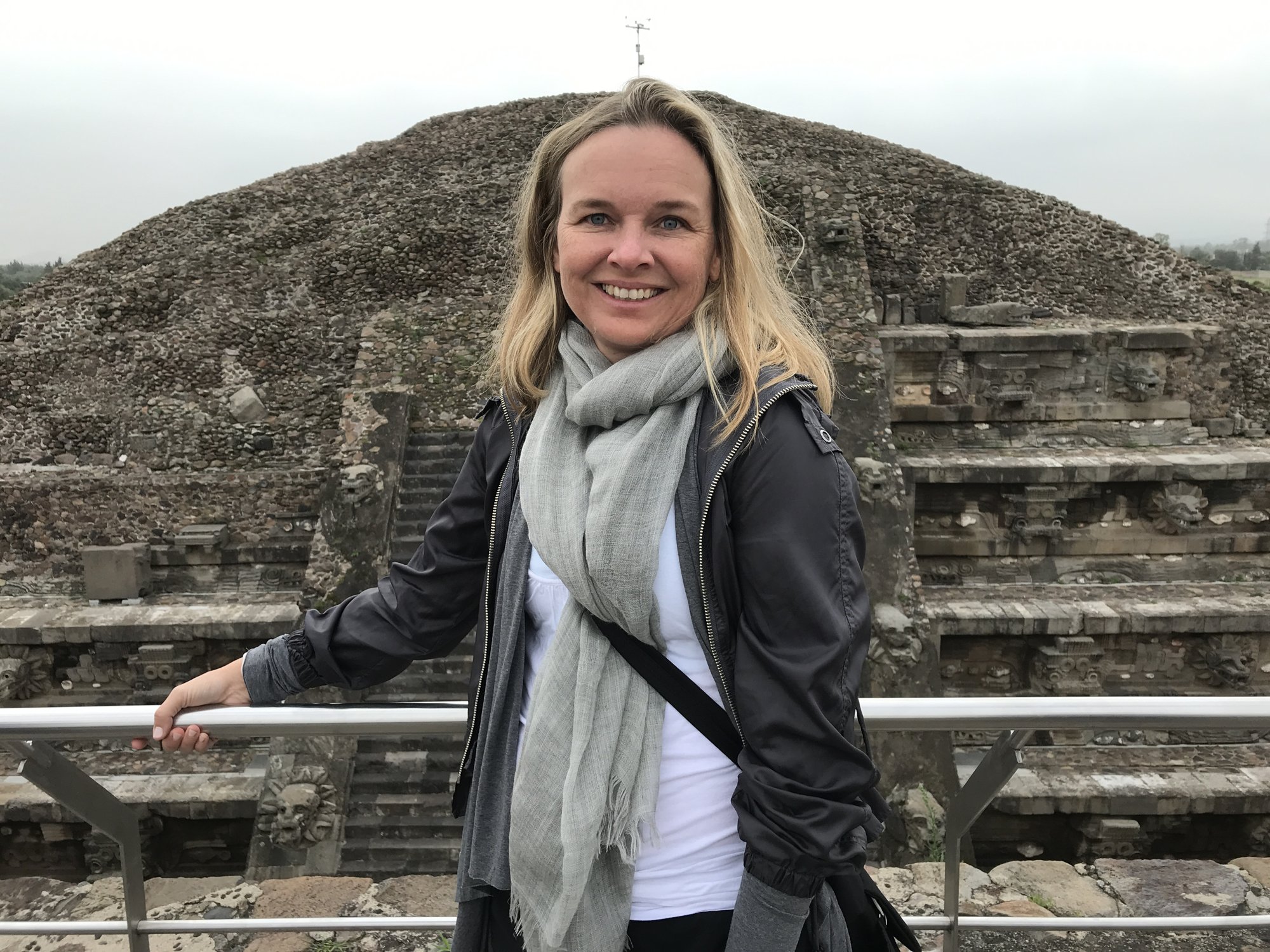She-Stache in a scarf and gray jacket on a terrace with the Pyramid of the Moon behind her