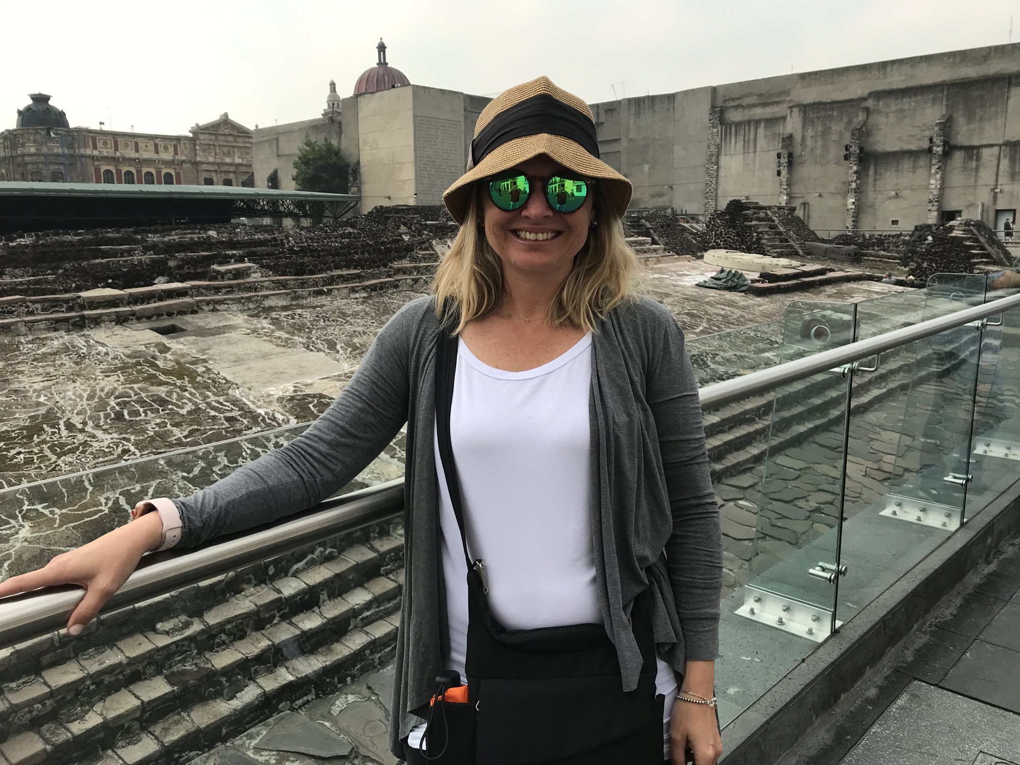 She-Stache in a sun hat and gray cardigan leaning on a railing above the excavated stone walls of Templo Mayor, with colonial buildings behind