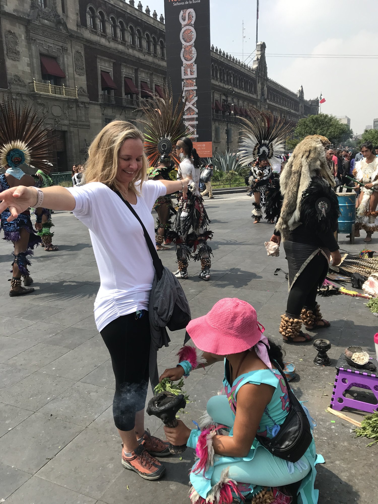 She-Stache with her arms out during a street cleansing ritual on the Zócalo, a healer in a pink hat waving smoking copal and herbs at her feet, dancers in feathered headdresses behind
