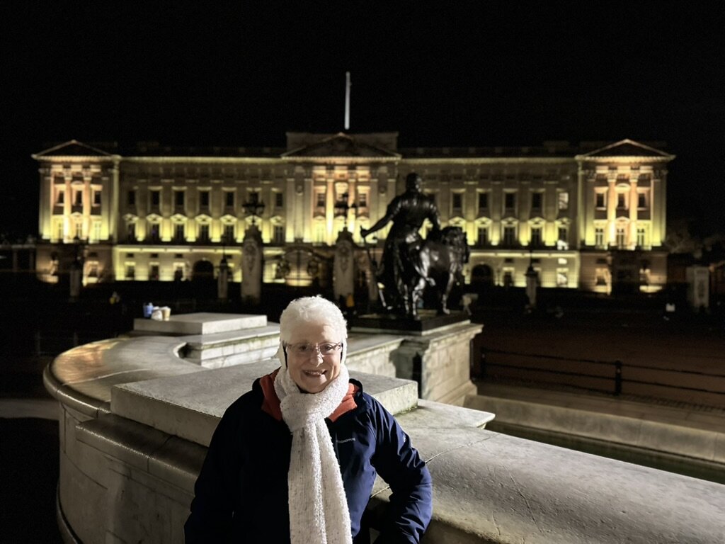 Mom smiling in front of Buckingham Palace lit up at night, with the Queen Victoria Memorial behind her