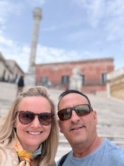 She-Stache and He-Stache smiling in front of the Roman column marking the end of the Appian Way at the top of the steps in Brindisi