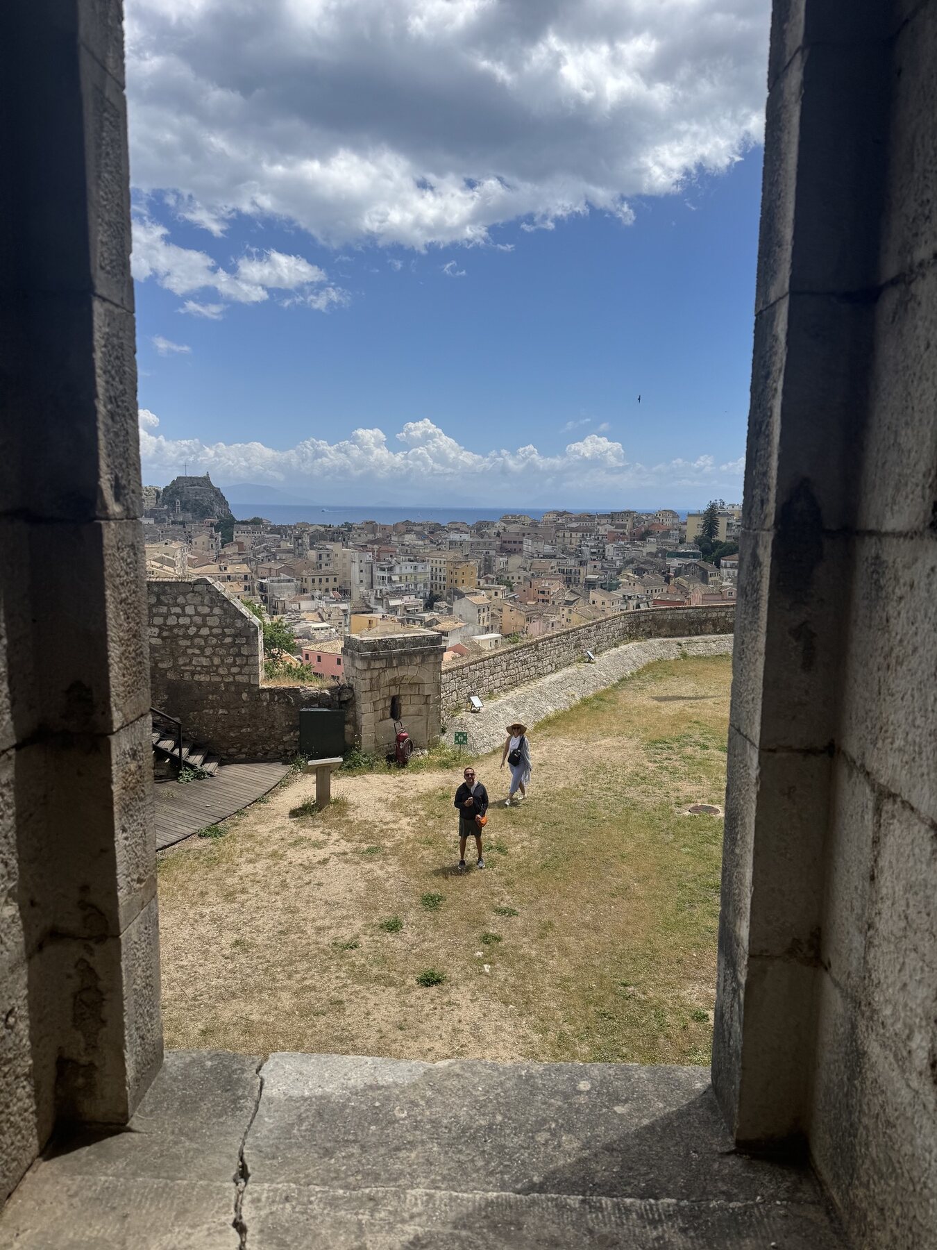 The Staches walking across a grassy fortress terrace, framed by a stone doorway, Corfu Town and the Old Fortress spread out below