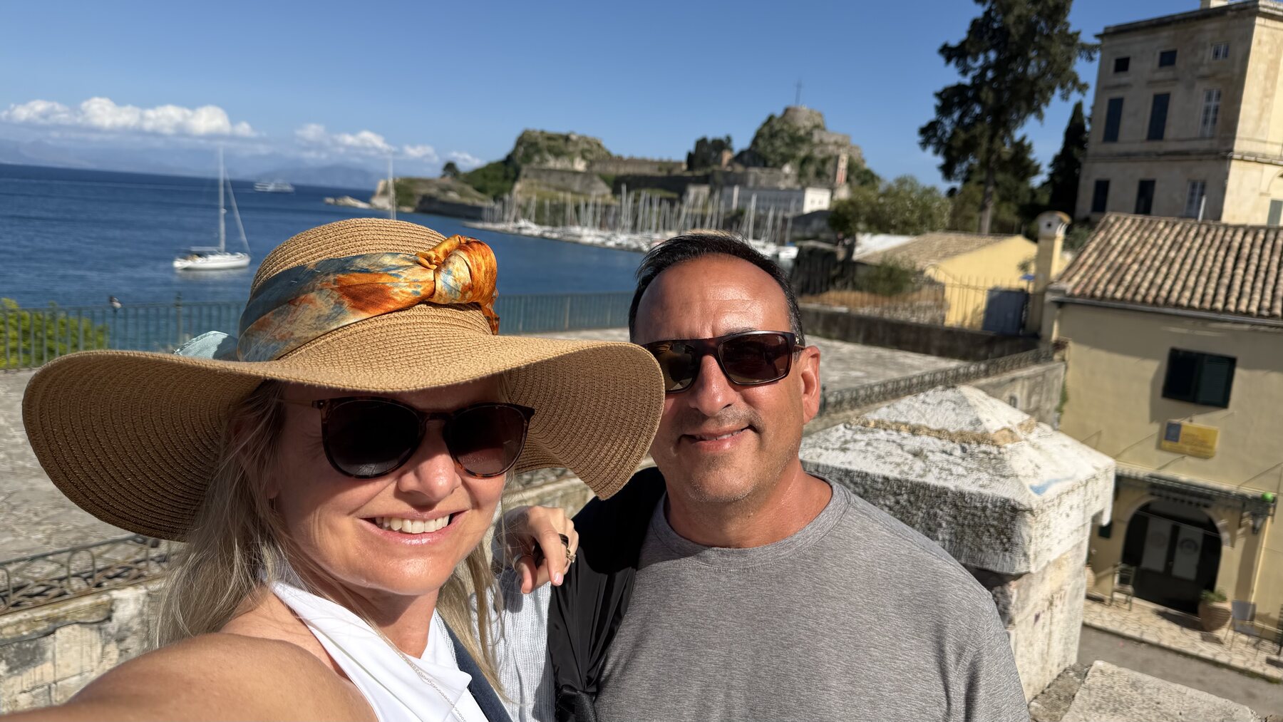 She-Stache in a wide straw hat and He-Stache smiling on the ramparts of Corfu, the Old Fortress and sailboats in the bay behind them