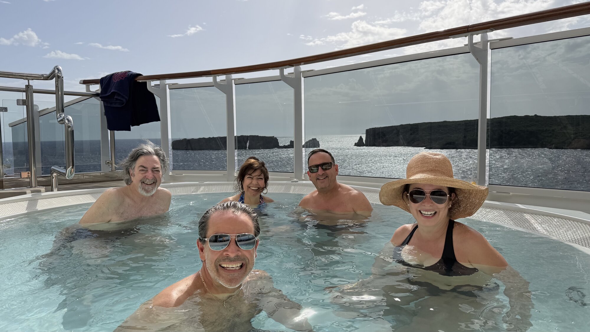 Five of our foodie-friend travel companions grinning in an Explora II hot tub, a rocky coastline behind them