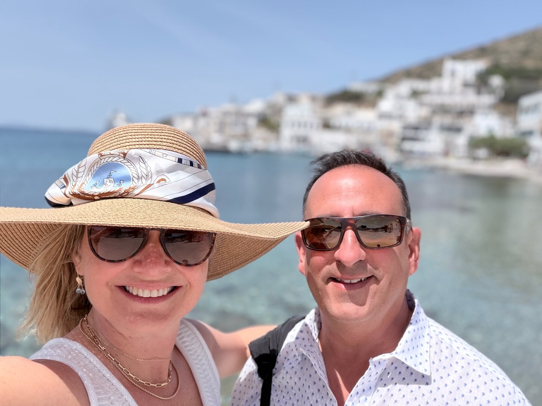 She-Stache in a wide straw hat and He-Stache smiling on the Katapola waterfront, whitewashed houses and clear water behind them