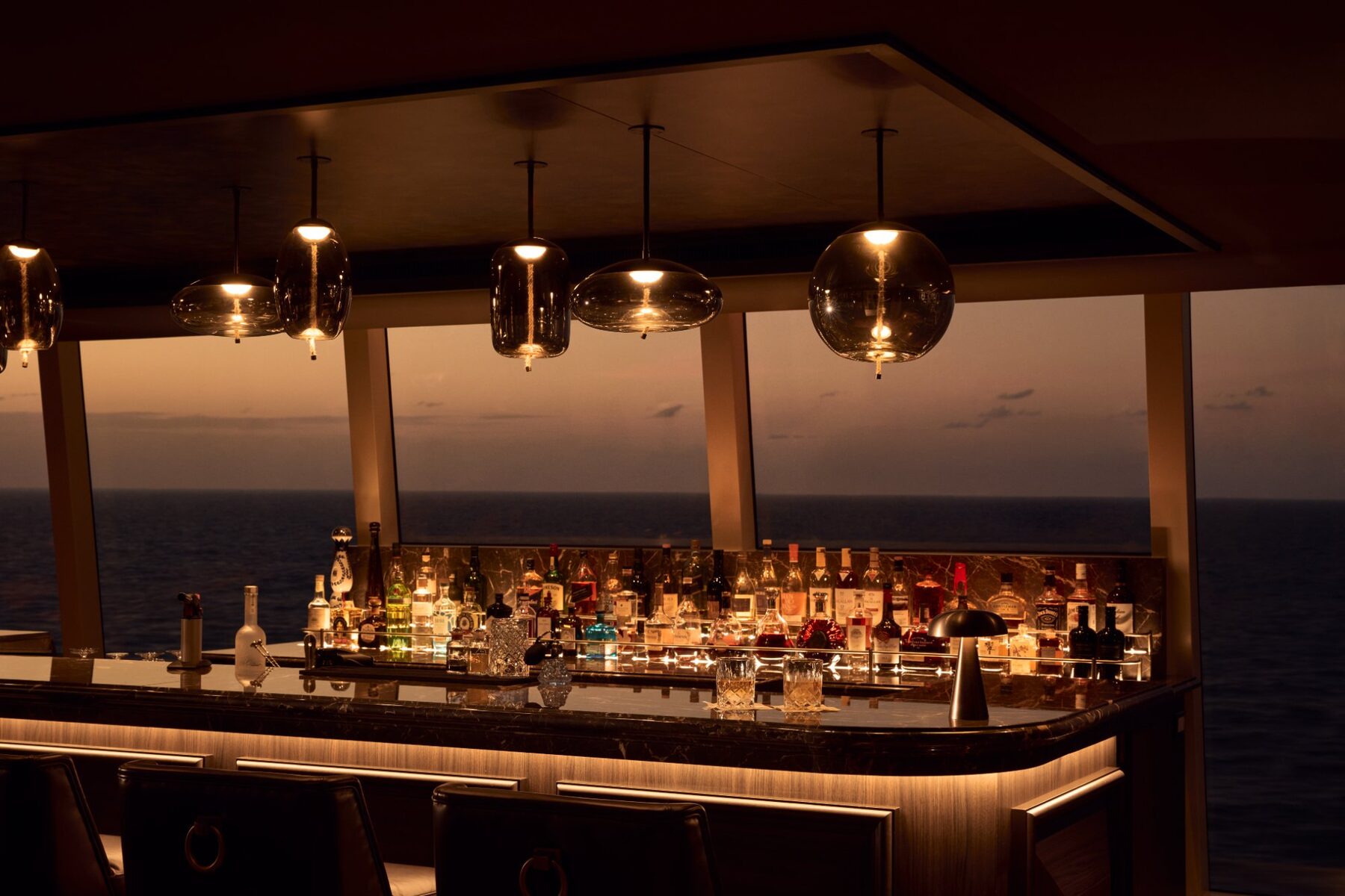 Explora media kit photo of the Malt Whisky Bar, a backlit wall of whiskies and spirits under glass pendant lights with the sea at sunset beyond