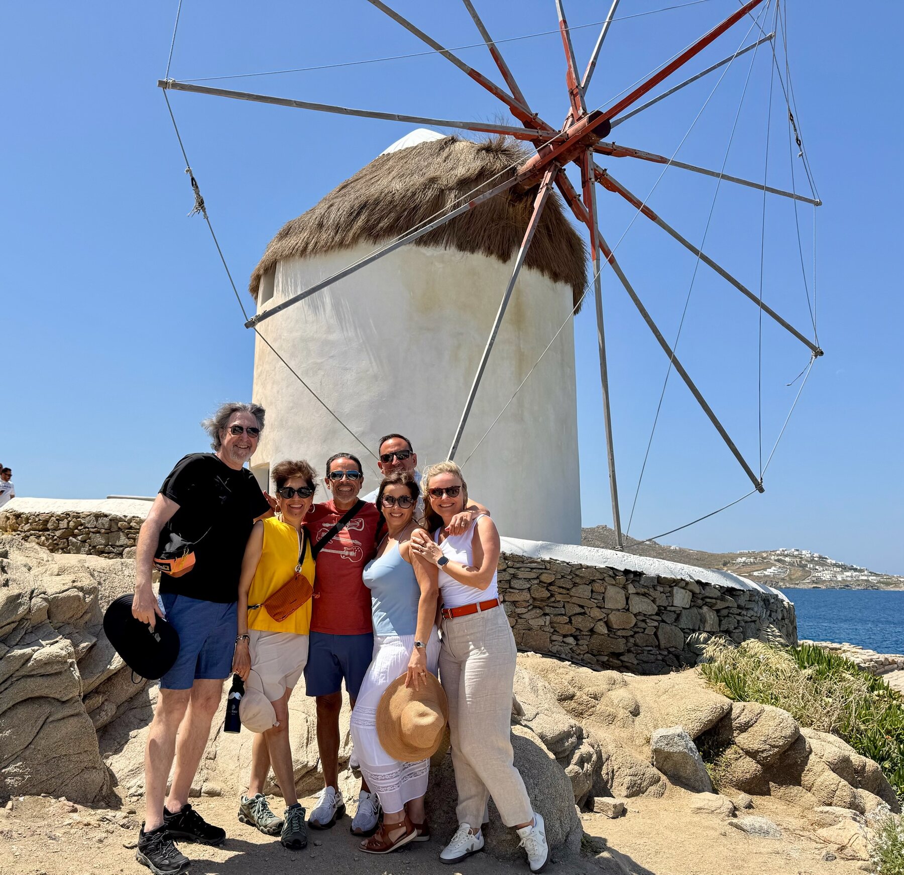 The Staches and the foodie friends grinning in front of a thatched Mykonos windmill, the sea and white hillside villas behind them
