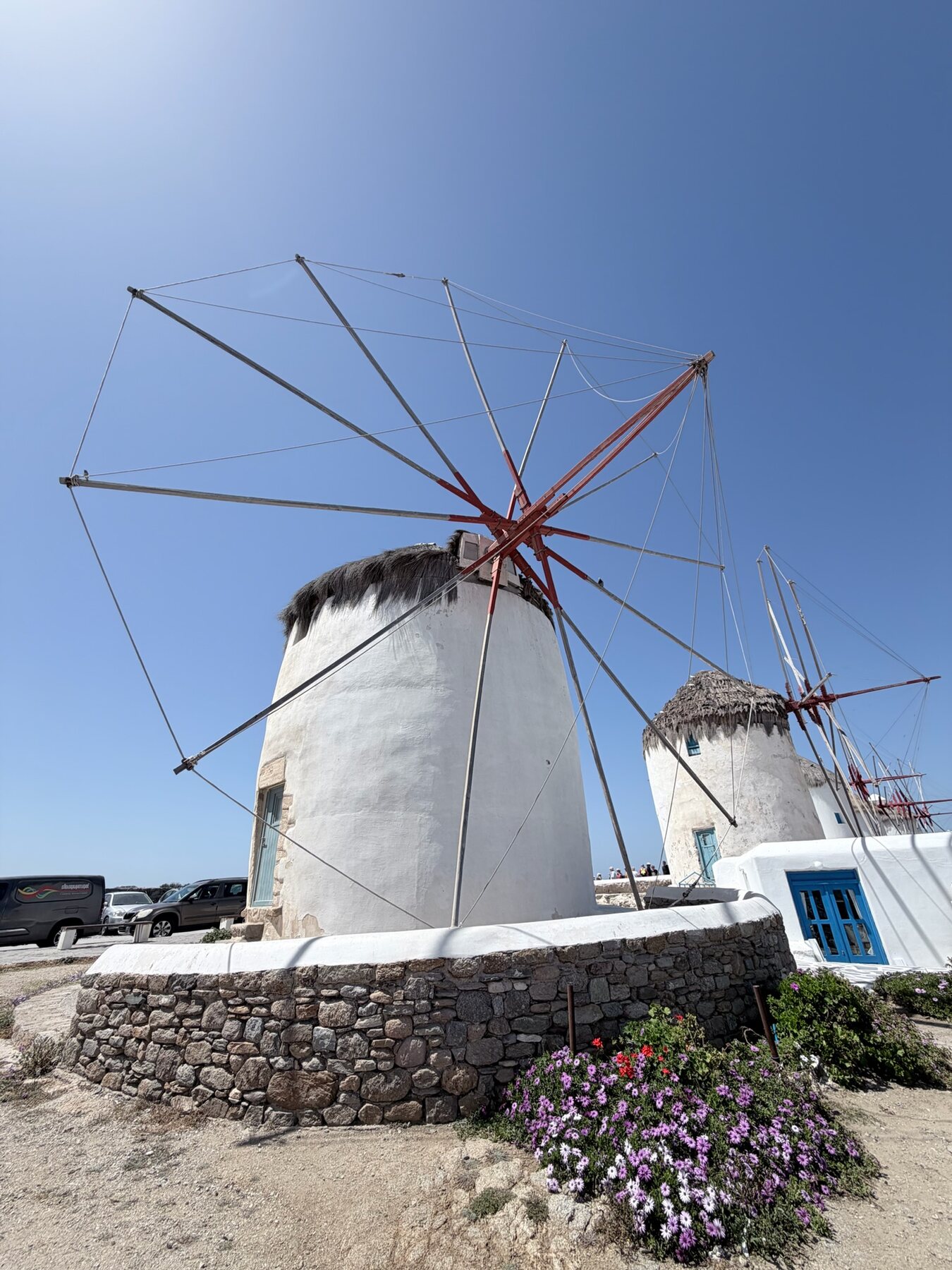 A whitewashed Mykonos windmill with red-and-white spokes against a deep blue sky, purple daisies at its stone base