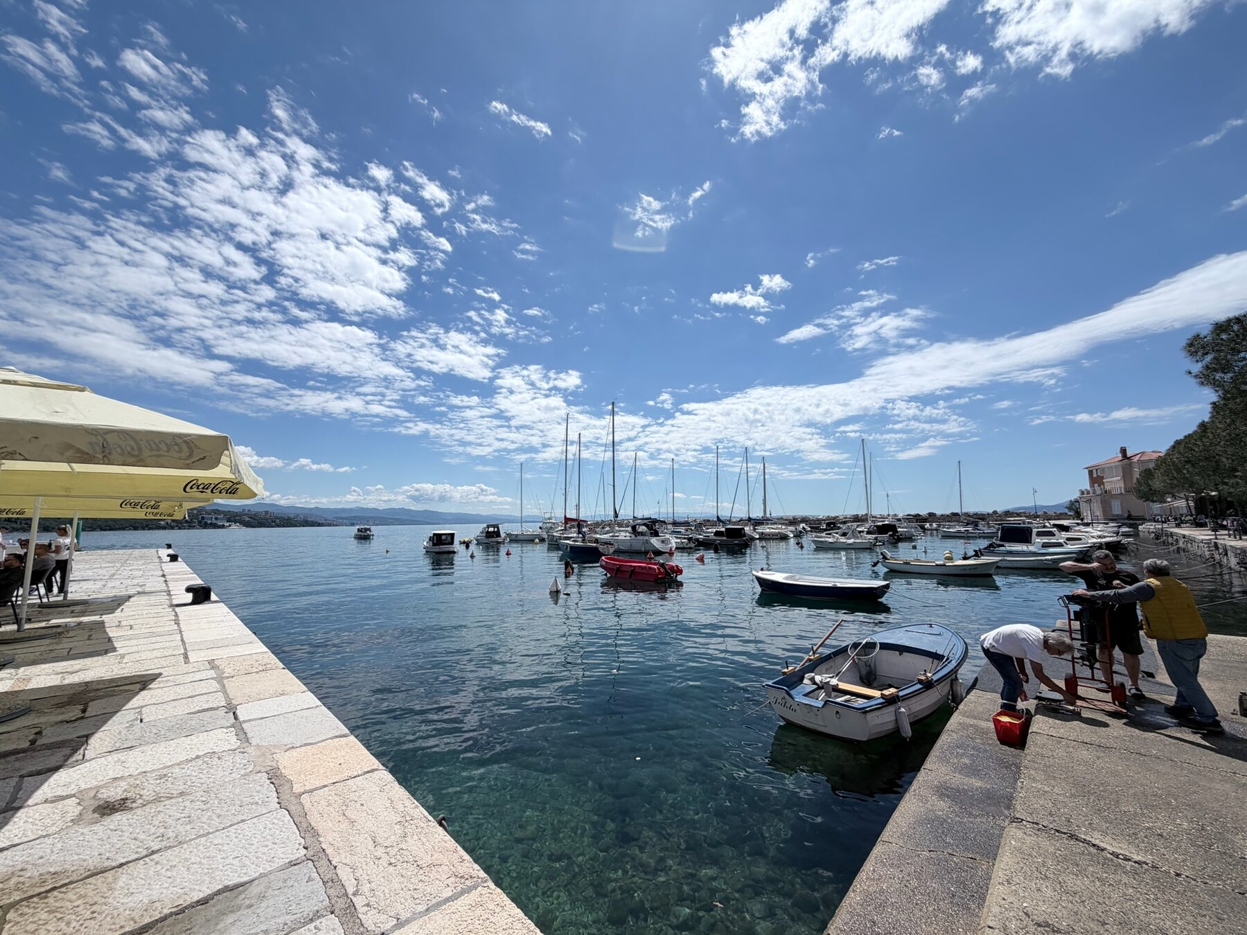 A small Opatija harbor with fishing boats and sailboats on clear turquoise water under a wide blue sky