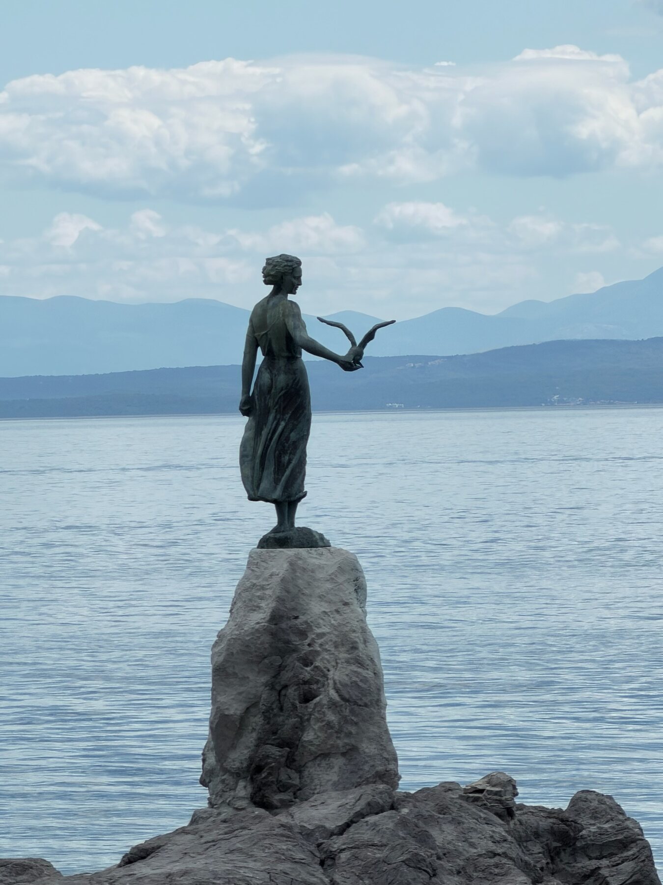 The bronze Maiden with the Seagull statue on a rock above the sea in Opatija, mountains across the bay