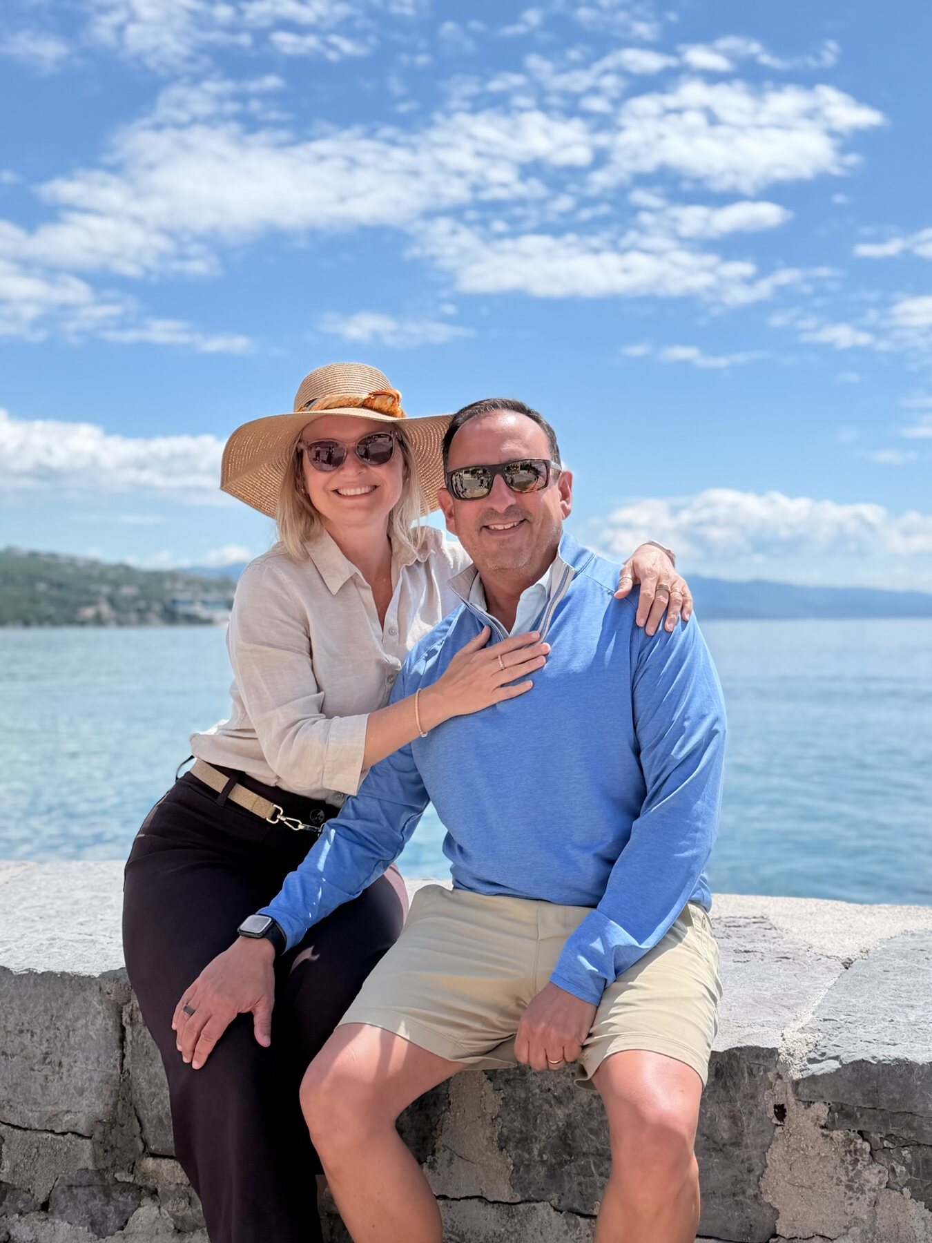 She-Stache in a wide straw hat with her arm around He-Stache, sitting on a stone seawall in Opatija with the blue Kvarner Bay behind them