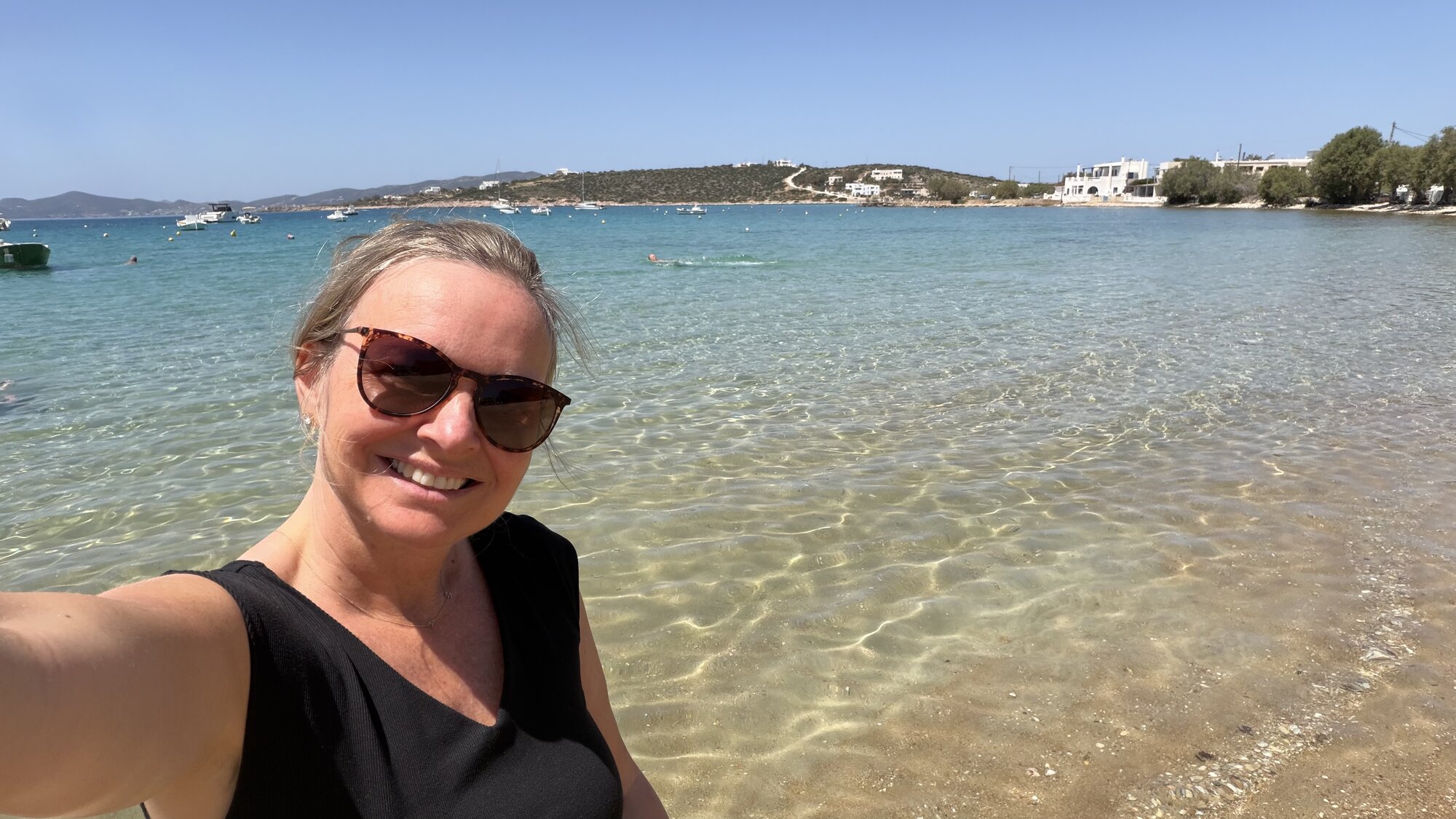 She-Stache in sunglasses taking a selfie at the edge of clear turquoise water on a Paros beach, white houses on the far shore