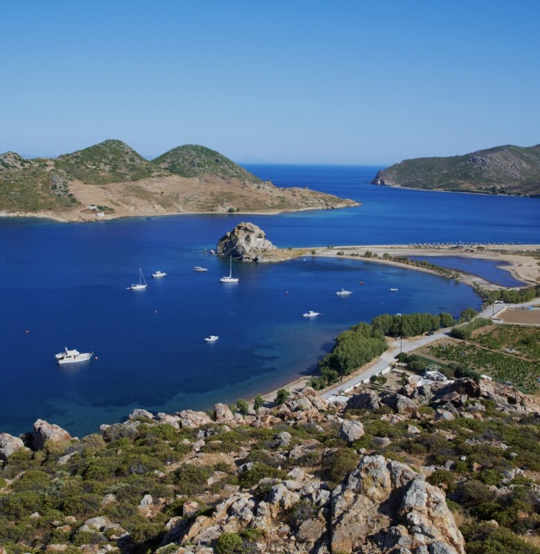 Stock photo of a deep blue Patmos bay dotted with sailboats, rocky green hills and a sandy spit beyond