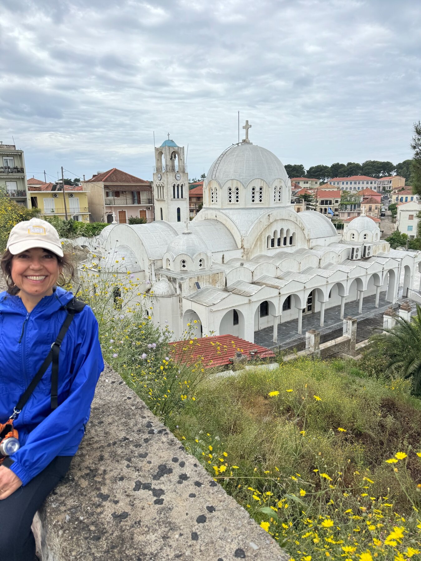 A travel companion in a blue rain jacket smiling on a stone wall above the white domed church of Pylos, red-roofed houses behind it