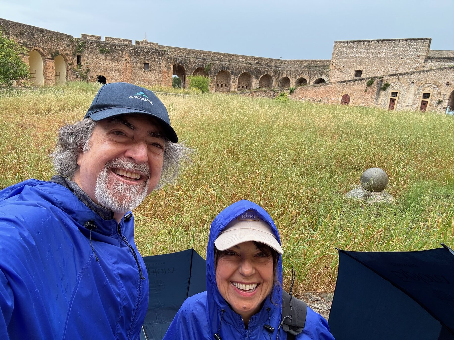 Two of our travel companions in matching blue rain jackets grinning in the grassy courtyard of the Niokastro fortress in Pylos, umbrellas at their feet