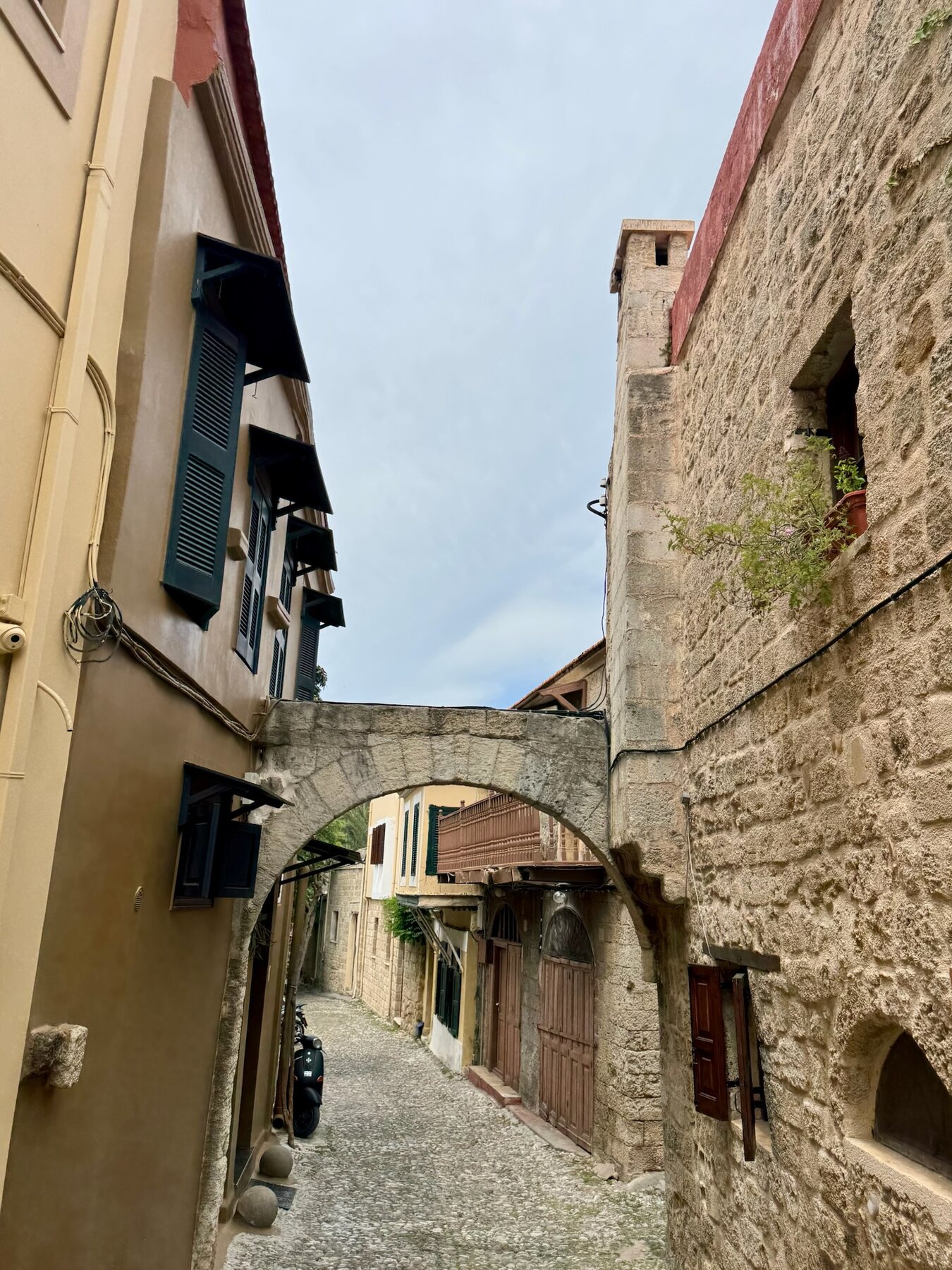 A cobbled lane in the old town of Rhodes passing under a stone arch between ochre houses with green shutters