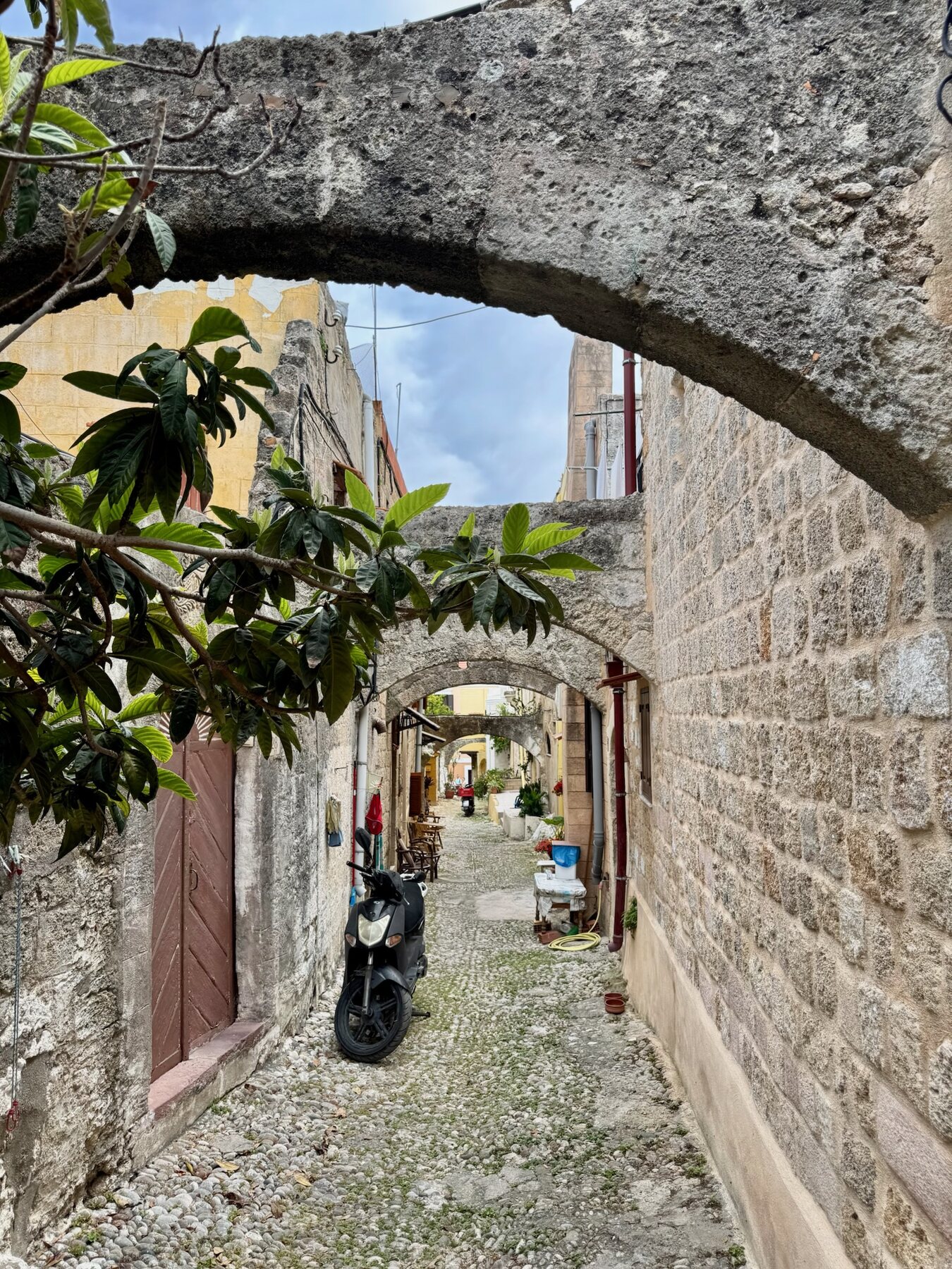 A narrow cobbled alley in Rhodes under a series of stone arches, a scooter parked beneath loquat leaves