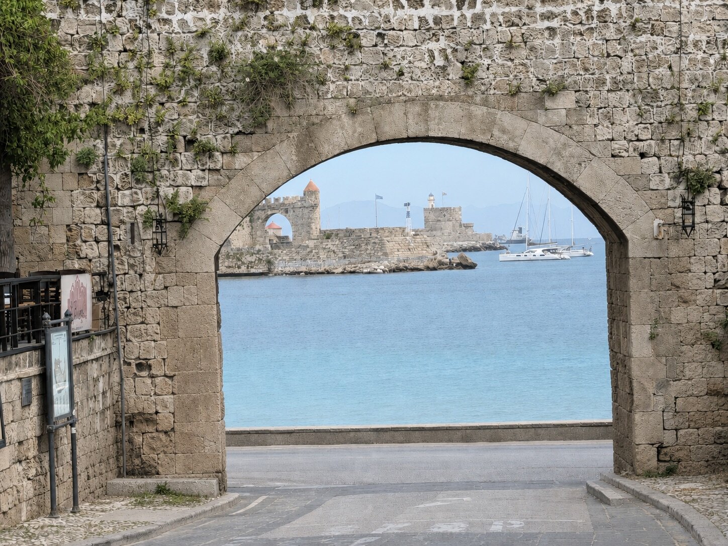 A stone gate in the medieval walls of Rhodes framing the turquoise harbor, the windmill fort and Saint Nicholas fortress across the water