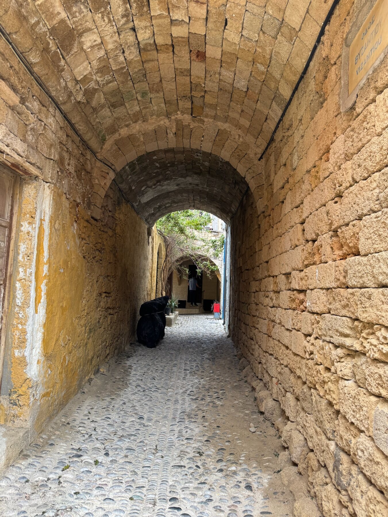 A vaulted stone passage with ochre walls opening onto a leafy courtyard in the Rhodes old town