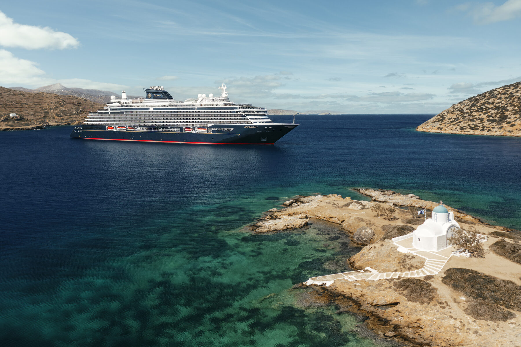 Explora media kit photo of Explora II at anchor in clear turquoise water off a rocky Greek island with a small white chapel
