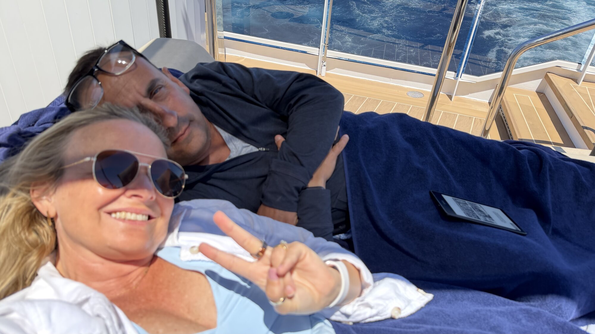 She-Stache flashing a peace sign on a shaded lounger aboard Explora II, with He-Stache napping beside her