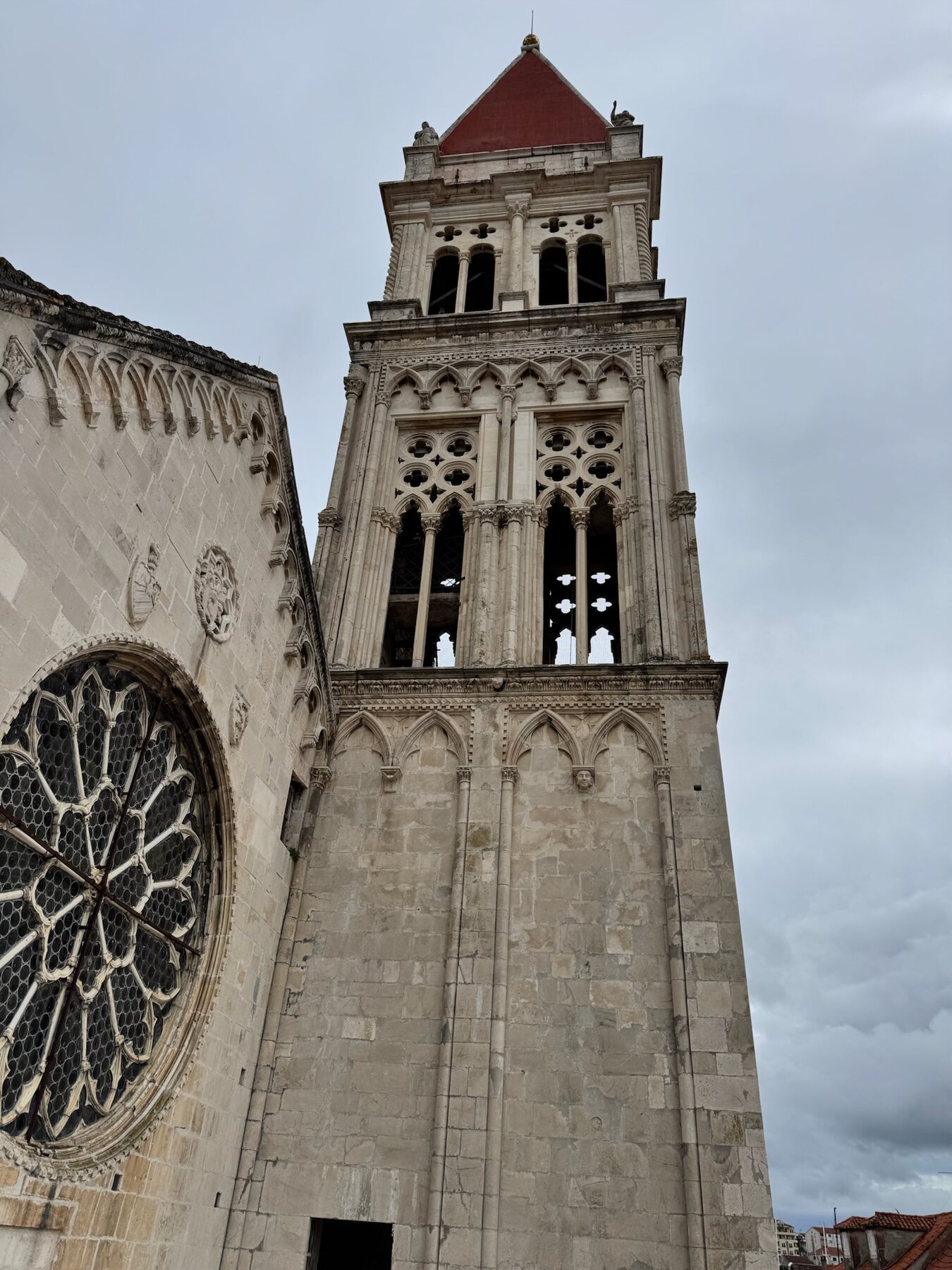 The tall Gothic bell tower of Trogir cathedral with a red pyramid roof beside a large rose window, under grey skies