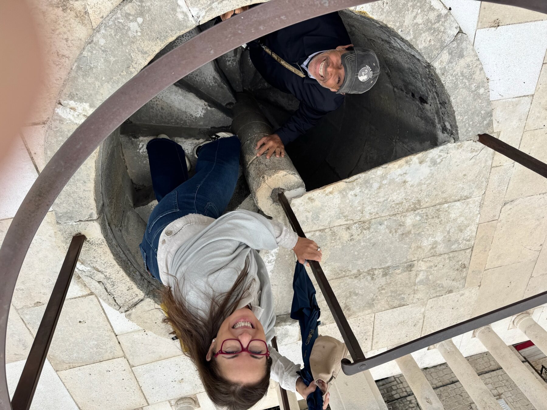Looking down a narrow stone spiral staircase inside the Trogir bell tower, two travel companions grinning up at the camera