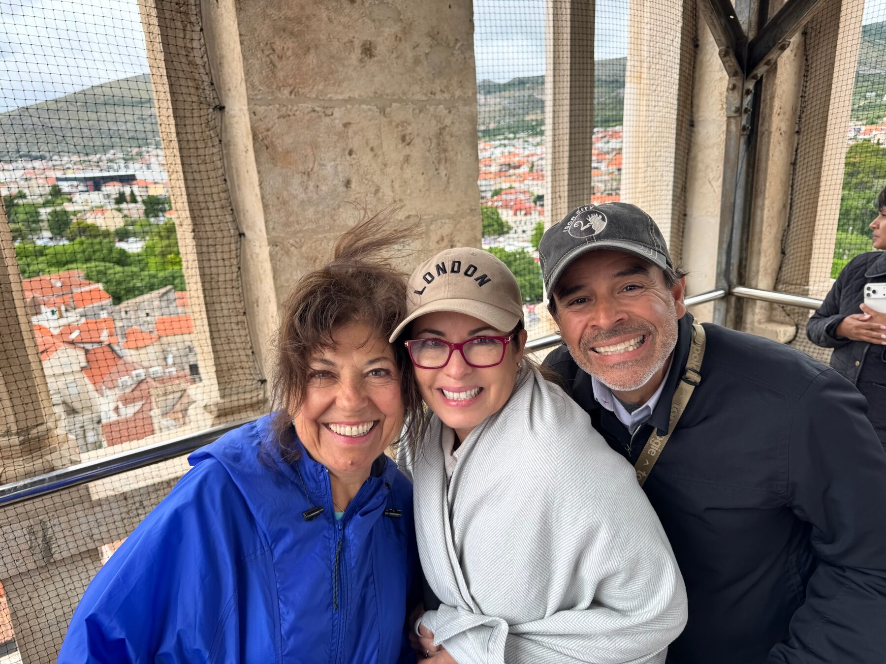 Three of our travel companions grinning and windswept at the top of the Trogir cathedral bell tower, red rooftops and green hills behind them