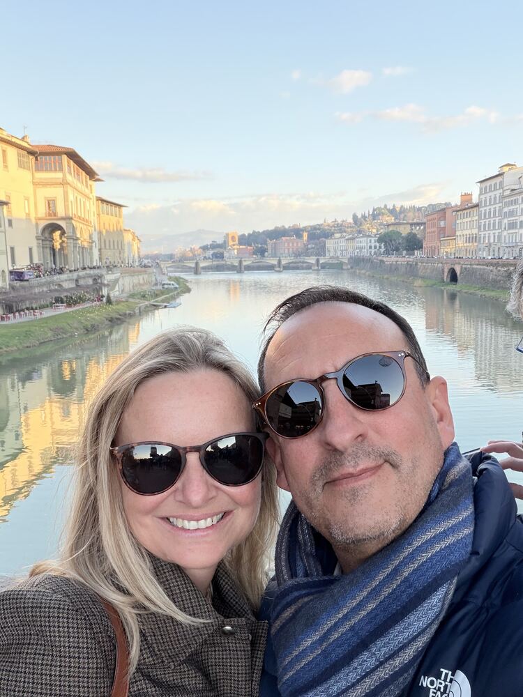 She-Stache and He-Stache in sunglasses and scarves on a bridge over a river lined with Italian buildings