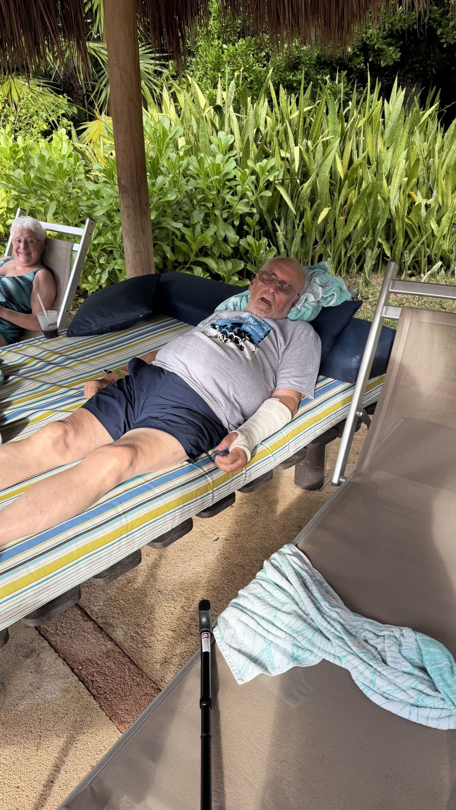 A man with his arm in a cast dozing on a striped lounger under a thatched palapa
