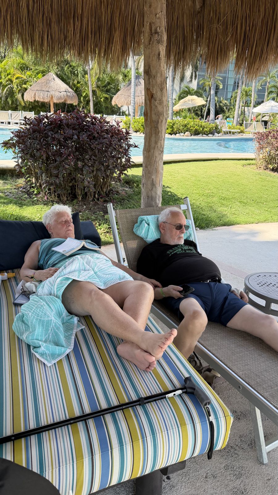 Two guests asleep on striped loungers under a palapa beside the pool