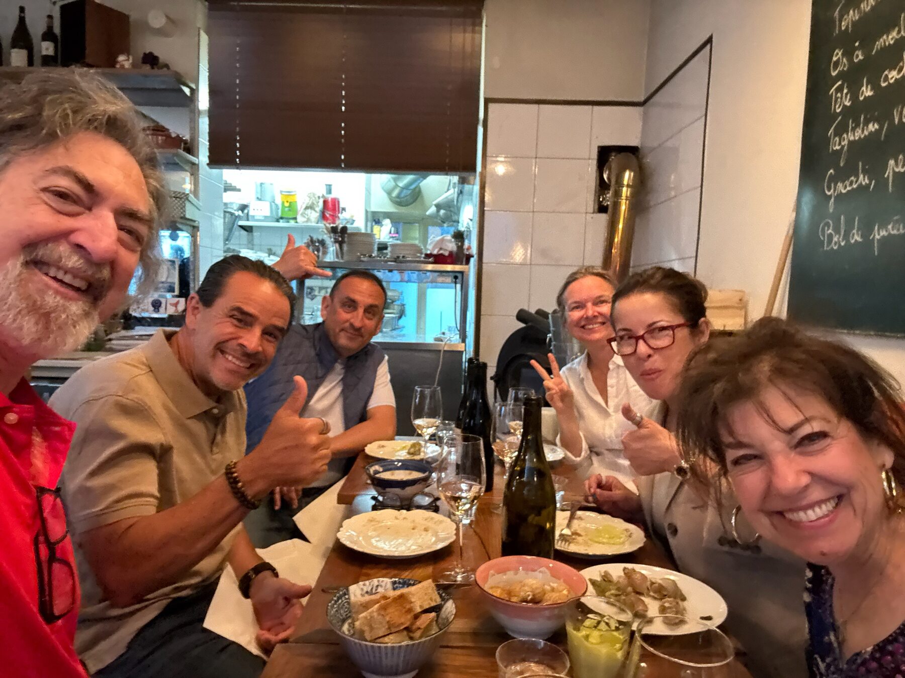 The group around a crowded bistro table with plates, bread and wine bottles, one friend giving a thumbs up