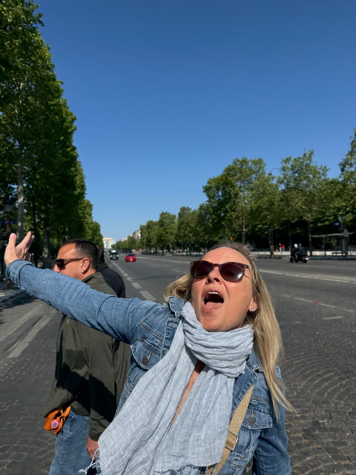 She-Stache throwing her arms wide in mock astonishment in the middle of a nearly empty Champs-Elysees