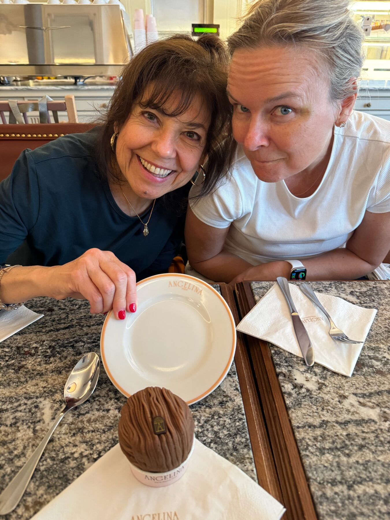 Two women grinning at a cafe table over a glossy chocolate dome pastry on a white plate