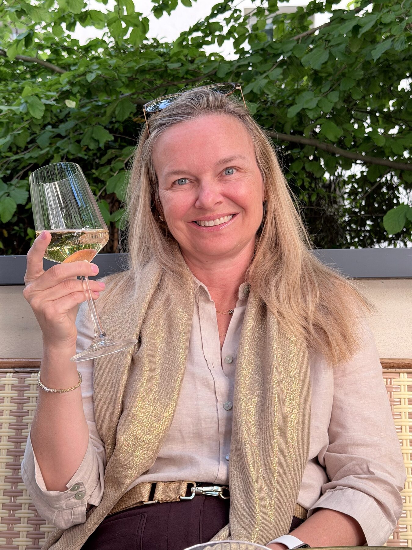 She-Stache smiling with a glass of white wine on a leafy Paris terrace