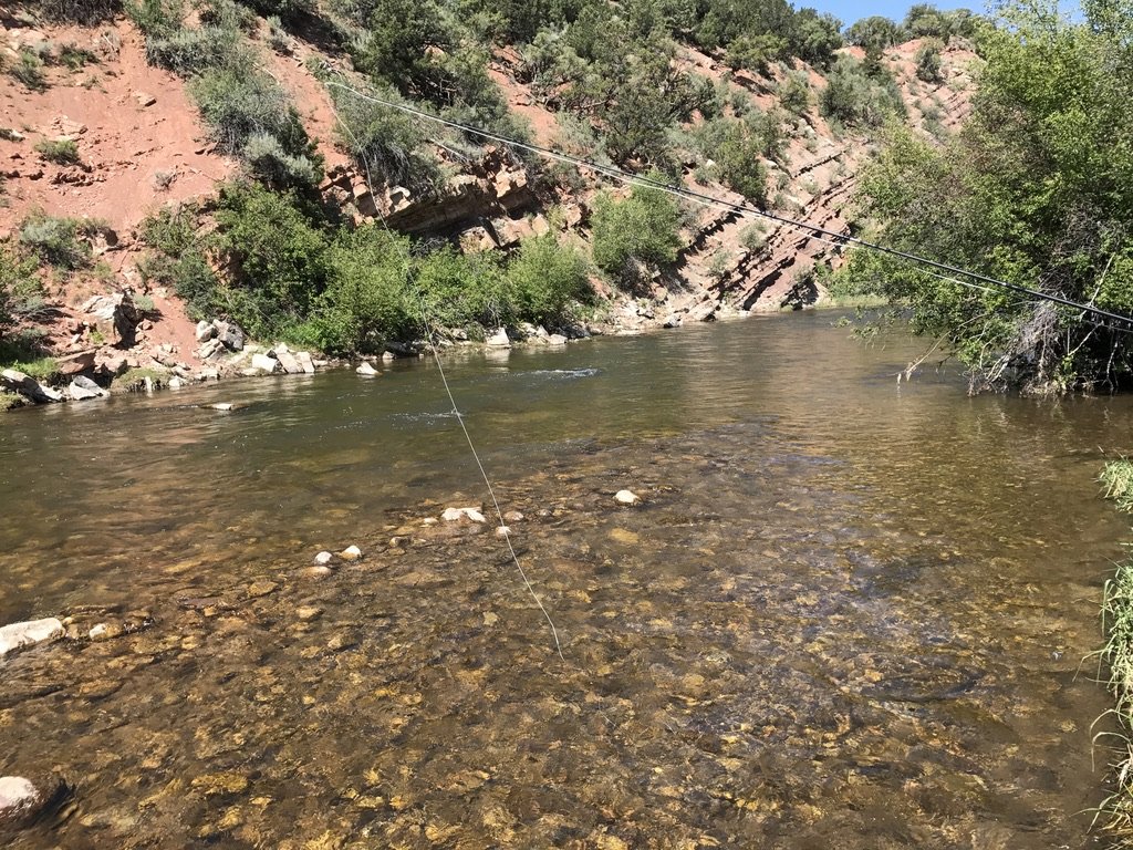 A fly line stretched across a clear, shallow river below red rock and green brush