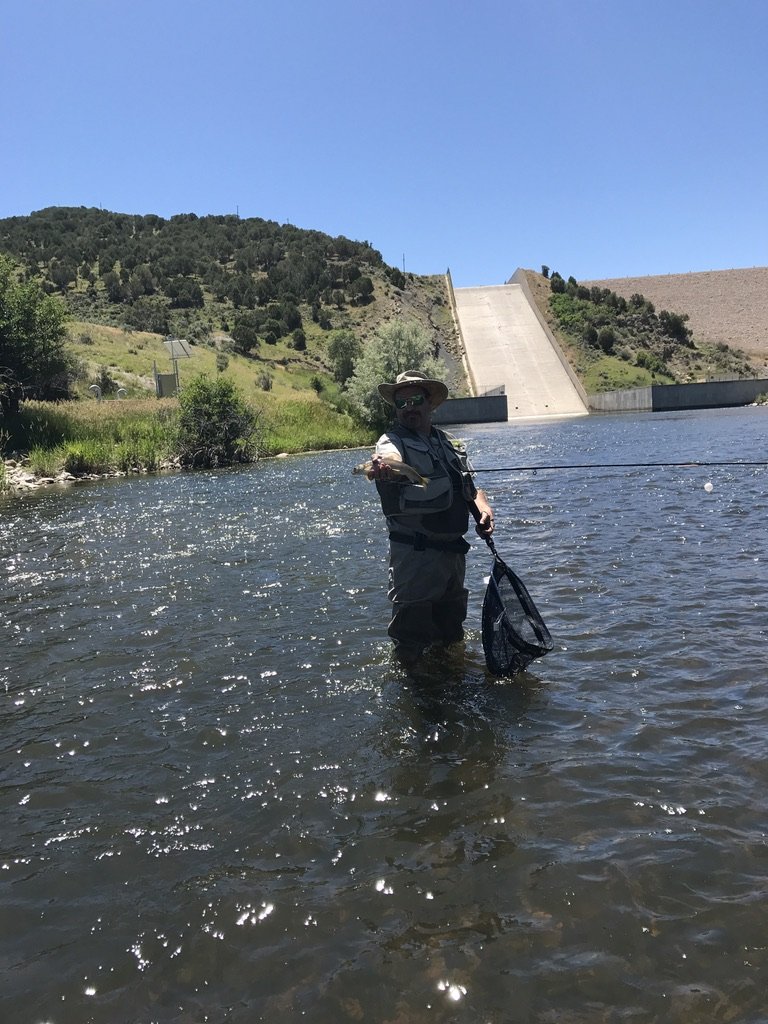 He-Stache in waders and a sun hat standing in the Weber River below the Rockport Reservoir spillway, holding up a trout with a net in his other hand