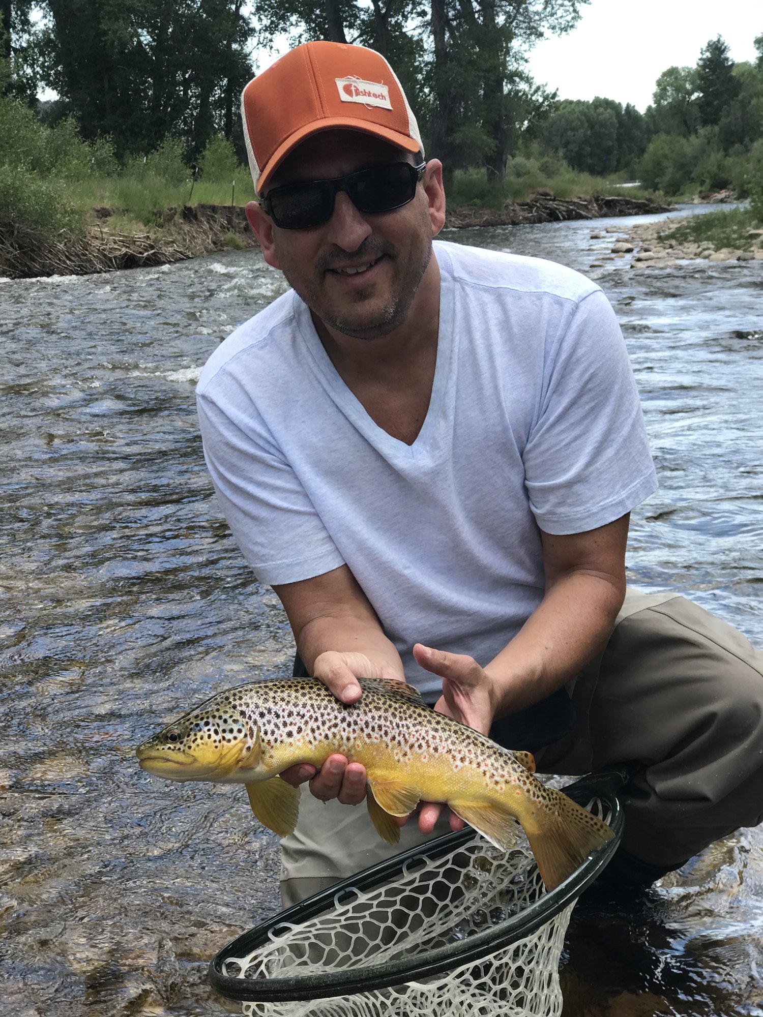 He-Stache crouching in a mountain stream, holding a brown trout above a landing net