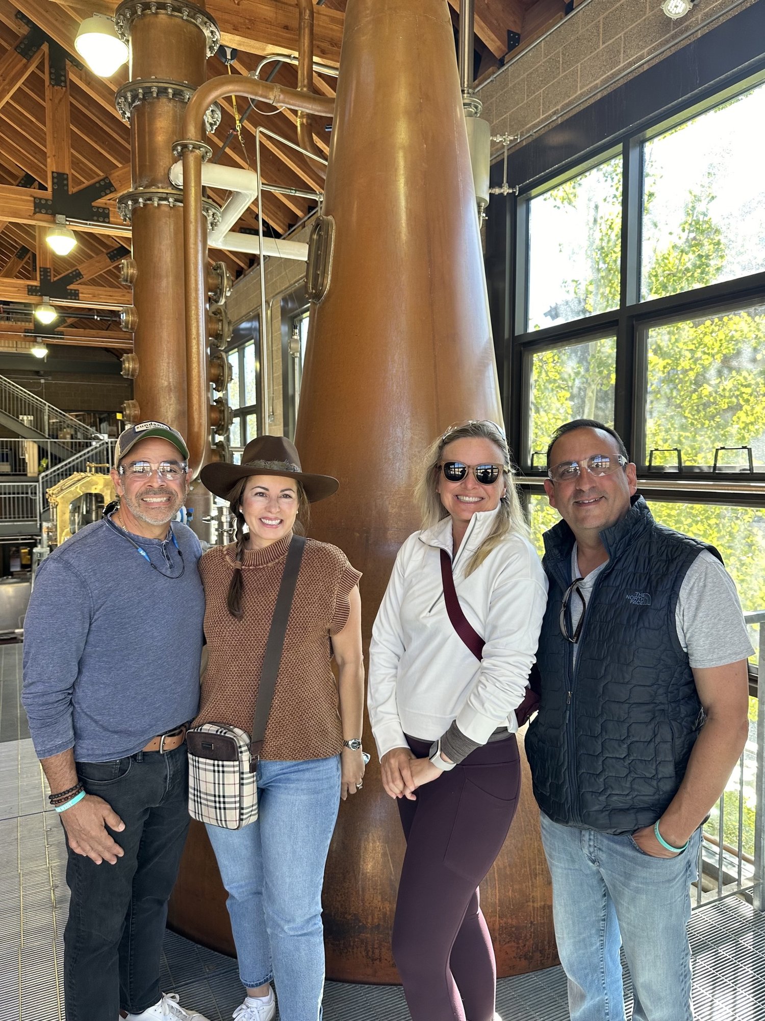 She-Stache, He-Stache and two friends in safety glasses standing in front of a tall copper still at High West&rsquo;s distillery