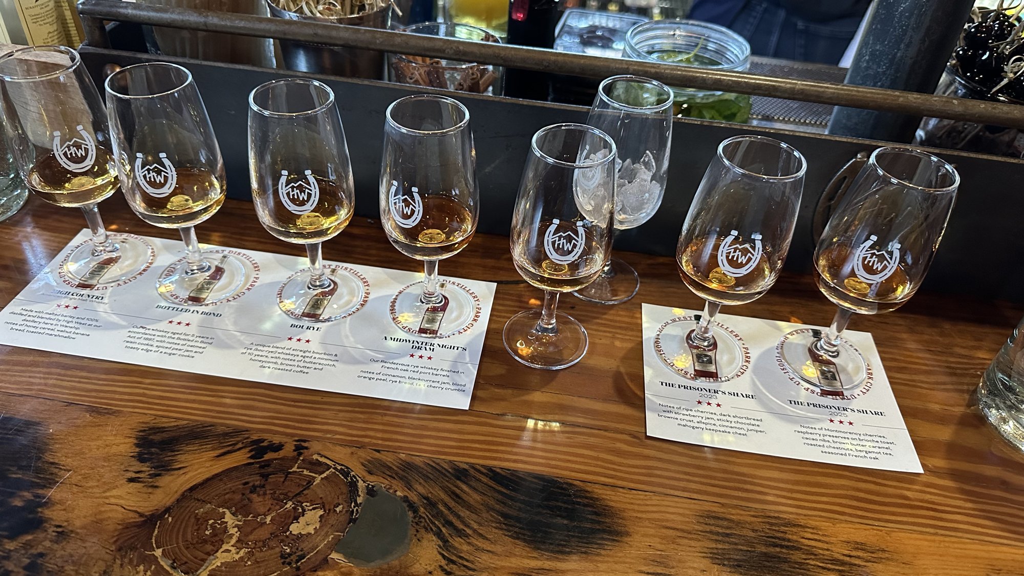 A row of seven High West tasting glasses of whiskey on printed tasting mats along a wooden bar