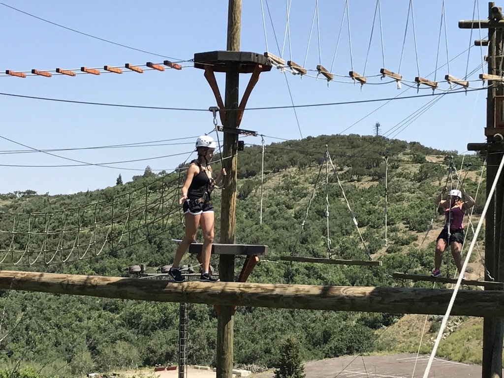 Two of the Wee-Staches in helmets and harnesses balancing on the high ropes course at Utah Olympic Park, with green hills behind