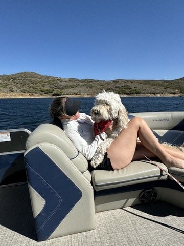 Pup-Stache sitting on the lap of a woman in a white sun shirt and visor on a pontoon boat, with the Jordanelle and dry hills behind them
